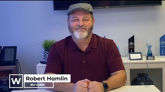 Man with gray beard and flat cap sitting at a desk with awards in the background, captioned Robert Hamlin, WLF client.