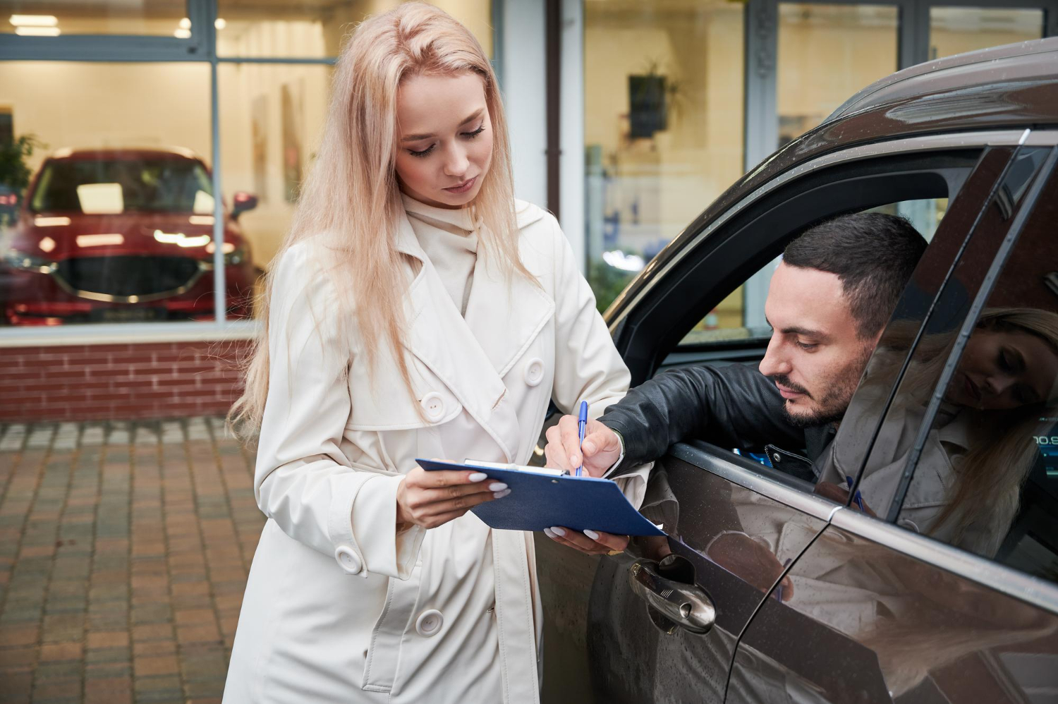 Man sitting in a car signing a document held by a woman standing outside wearing a white coat.